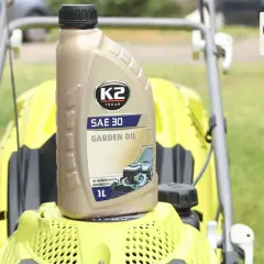 Butelka oleju K2 SAE 30 Garden Oil do kosiarki spalinowej, stojąca na żółtej obudowie maszyny.