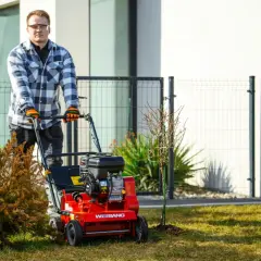 Mężczyzna w okularach i kratę prowadzi czerwony wertykulator, który robi nacięcia w trawniku, przygotowując go do wiosennych prac.