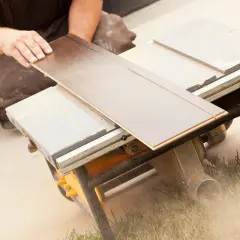 Ręce układają panel podłogowy na pile stołowej, która jest idealną tarczą do cięcia paneli podłogowych. W tle widać kurz.