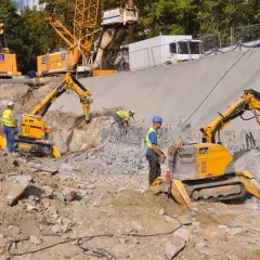 Na placu budowy pracują roboty wyburzeniowe, które kruszą beton. W tle widać koparki i dźwigi.