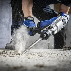 Mocny udar w wiertarce kruszy beton, wzbijając tumany pyłu. Pracownik w niebieskich rękawicach pewnie trzyma narzędzie.
