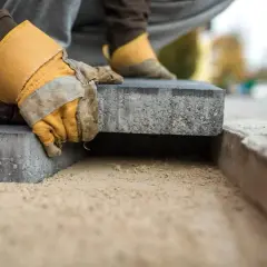 Dłoń w rękawicy układa kostkę brukową na piasku, przygotowując podłoże do utwardzania gruntu.