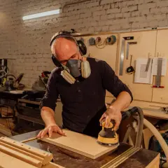 Mężczyzna w masce i słuchawkach szlifuje drewno szlifierką kątową.
