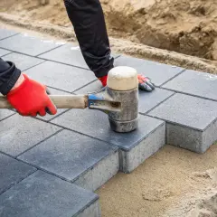 Ręce w czerwonych rękawiczkach używają gumowego młotka jako układarka do kostki, precyzyjnie dopasowując szare płyty.