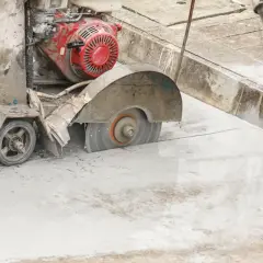 Maszyna do cięcia betonu pracuje, tworząc pył. Robotnik w czerwonej rękawicy niesie wiadro.