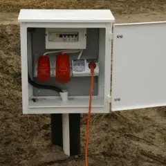 Tymczasowe przyłącze na czas budowy: biała skrzynka z bezpiecznikami, dwoma czerwonymi gniazdami siłowymi i jednym gniazdem elektrycznym, z pomarańczowym kablem.