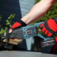 Ręce w czerwonych rękawicach trzymają piłę szablastą Tryton. To nie Black+Decker, ale równie solidne narzędzie do cięcia.