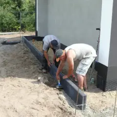 Dwóch mężczyzn układa krawężniki, jeden używa młotka do zagęszczania betonu.