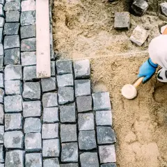 Ręczne układanie kostki brukowej, pracownik w niebieskich rękawiczkach ubija kamienie gumowym młotkiem.