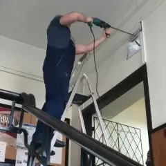 Mężczyzna w ogrodniczkach stoi na drabinie, wiercąc w ścianie. Trwa budowa drabiny, która ułatwia mu pracę.