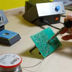 Ręka trzyma lutownicę, która służy do łączenia elementów na płytce drukowanej. Obok stojak na lutownicę i rolka cyny.