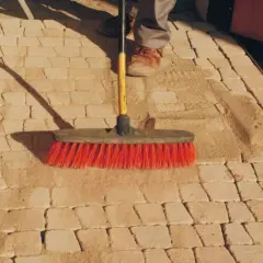 Szczotka z czerwonym włosiem usuwa piasek z kamiennej ścieżki, pokazując, jak usunąć igliwie z kamieni.