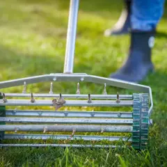 Aerator do trawy z kolcami na zielonej trawie. W tle widać nogi w gumowcach.