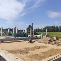 Budowa domu w toku. Betonowe słupy stoją na fundamencie, a w tle widać maszyny do zagęszczania gruntu i stosy piasku.