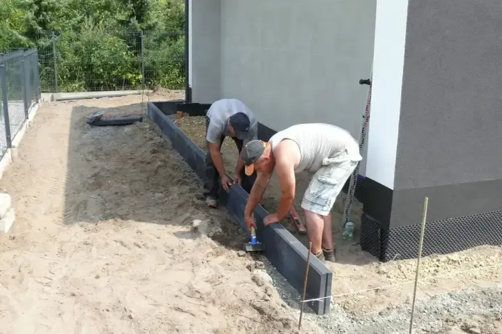 Dwóch mężczyzn układa krawężniki, jeden używa młotka do zagęszczania betonu.