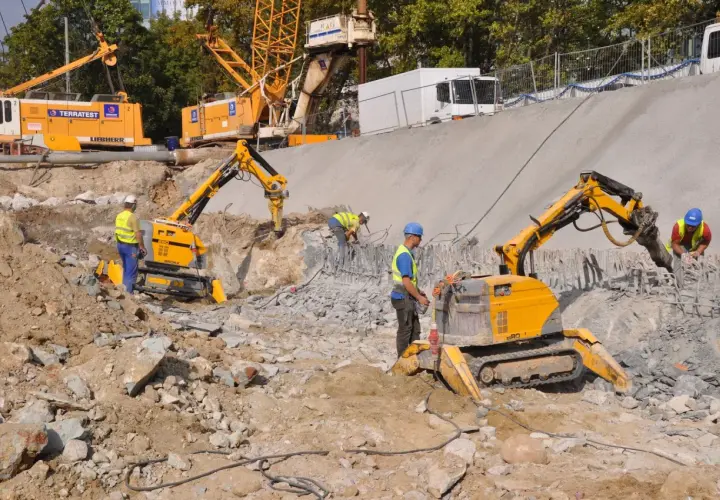 Na placu budowy pracują roboty wyburzeniowe, które kruszą beton. W tle widać koparki i dźwigi.