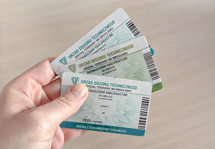 Ręka trzyma kilka zaświadczeń kwalifikacyjnych Urzędu Dozoru Technicznego, potwierdzających uprawnienia na zwyżki.