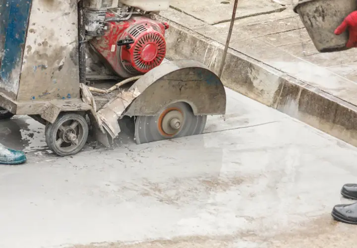 Maszyna do cięcia betonu pracuje, tworząc pył. Robotnik w czerwonej rękawicy niesie wiadro.