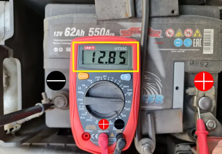 Multimetr pokazuje 12.85V, co jest wynikiem pomiaru napięcia elektrycznego akumulatora samochodowego.