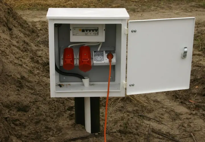 Tymczasowe przyłącze na czas budowy: biała skrzynka z bezpiecznikami, dwoma czerwonymi gniazdami siłowymi i jednym gniazdem elektrycznym, z pomarańczowym kablem.