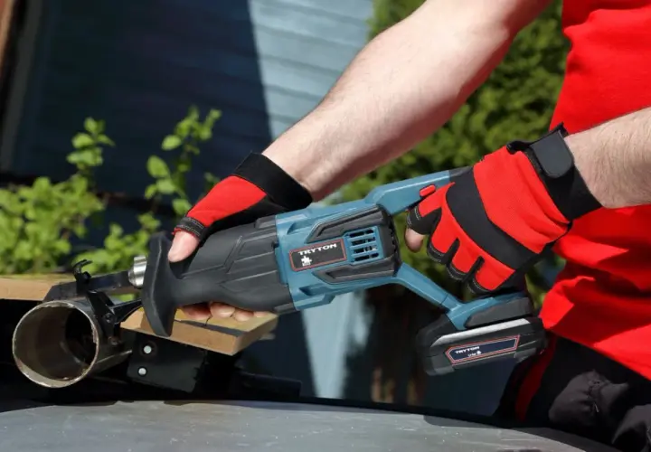 Ręce w czerwonych rękawicach trzymają piłę szablastą Tryton. To nie Black+Decker, ale równie solidne narzędzie do cięcia.