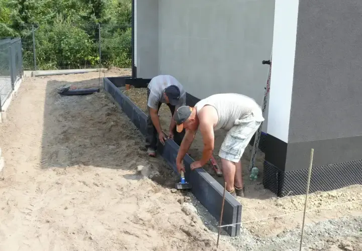 Dwóch mężczyzn układa krawężniki, jeden używa młotka do zagęszczania betonu.