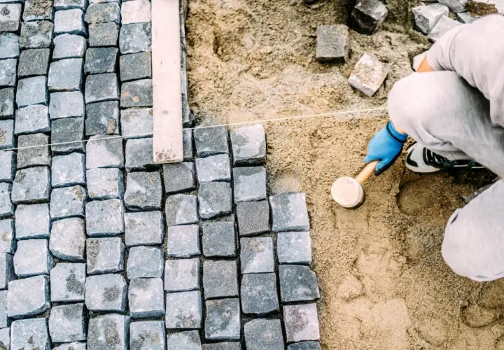 Ręczne układanie kostki brukowej, pracownik w niebieskich rękawiczkach ubija kamienie gumowym młotkiem.