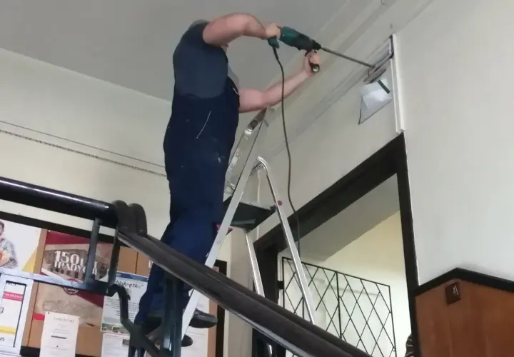 Mężczyzna w ogrodniczkach stoi na drabinie, wiercąc w ścianie. Trwa budowa drabiny, która ułatwia mu pracę.