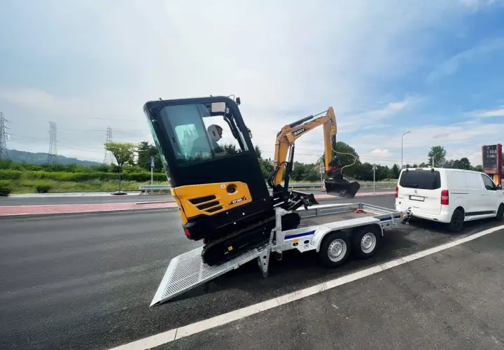 Mini koparka Sany SY18C na przyczepie, gotowa do transportu. Obok biały bus i inne przyczepy.