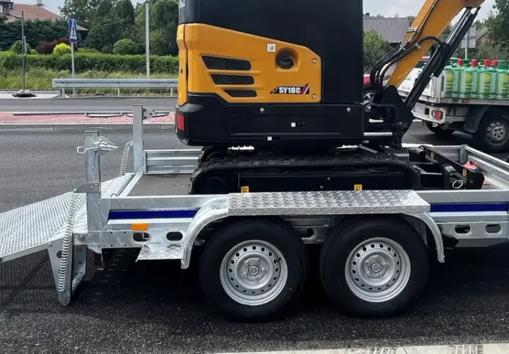 Żółta minikoparka na przyczepie, gotowa do transportu. Jej waga jest znacząca, ale przyczepa jest solidna.