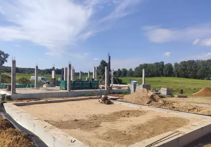 Budowa domu w toku. Betonowe słupy stoją na fundamencie, a w tle widać maszyny do zagęszczania gruntu i stosy piasku.