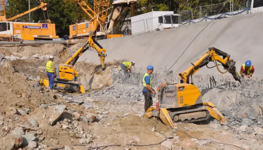 Na placu budowy pracują roboty wyburzeniowe, które kruszą beton. W tle widać koparki i dźwigi.