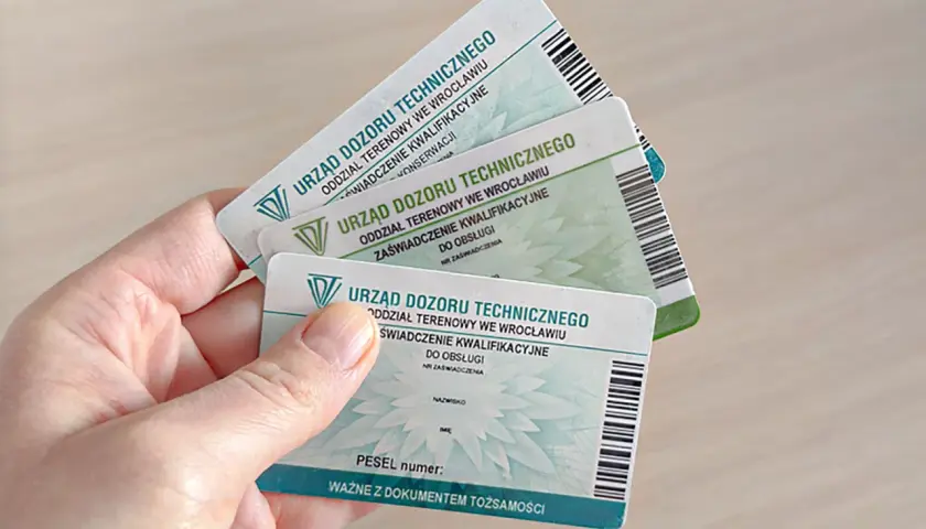 Ręka trzyma kilka zaświadczeń kwalifikacyjnych Urzędu Dozoru Technicznego, potwierdzających uprawnienia na zwyżki.