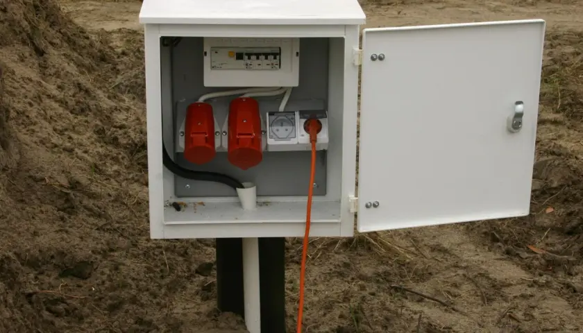 Tymczasowe przyłącze na czas budowy: biała skrzynka z bezpiecznikami, dwoma czerwonymi gniazdami siłowymi i jednym gniazdem elektrycznym, z pomarańczowym kablem.