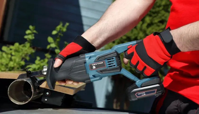 Ręce w czerwonych rękawicach trzymają piłę szablastą Tryton. To nie Black+Decker, ale równie solidne narzędzie do cięcia.