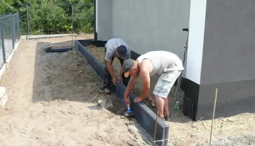 Dwóch mężczyzn układa krawężniki, jeden używa młotka do zagęszczania betonu.
