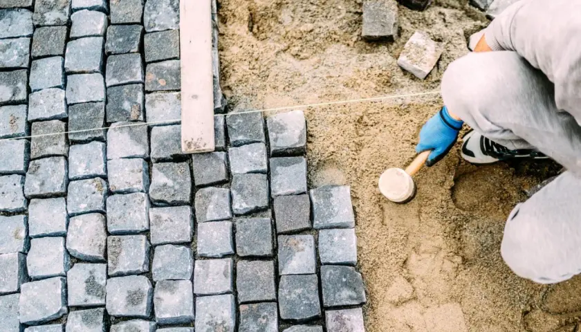 Ręczne układanie kostki brukowej, pracownik w niebieskich rękawiczkach ubija kamienie gumowym młotkiem.