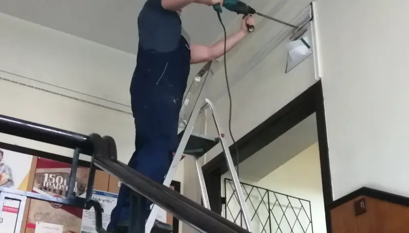 Mężczyzna w ogrodniczkach stoi na drabinie, wiercąc w ścianie. Trwa budowa drabiny, która ułatwia mu pracę.