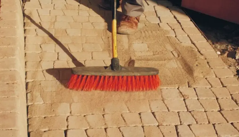 Szczotka z czerwonym włosiem usuwa piasek z kamiennej ścieżki, pokazując, jak usunąć igliwie z kamieni.