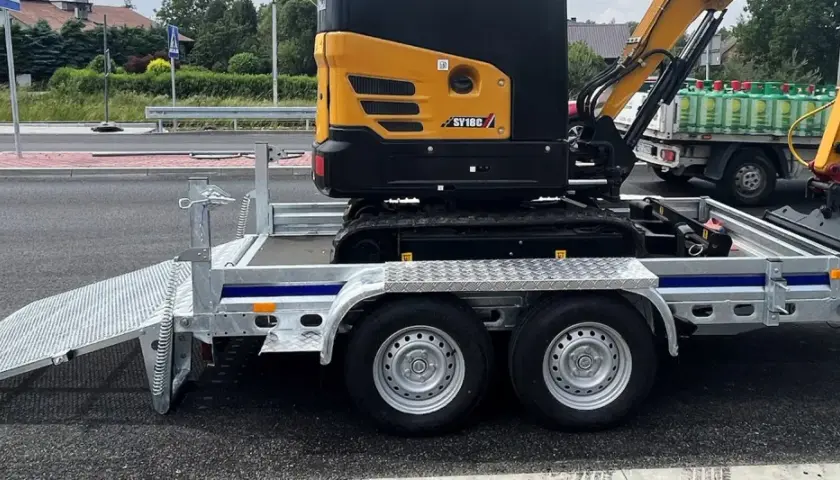 Żółta minikoparka na przyczepie, gotowa do transportu. Jej waga jest znacząca, ale przyczepa jest solidna.