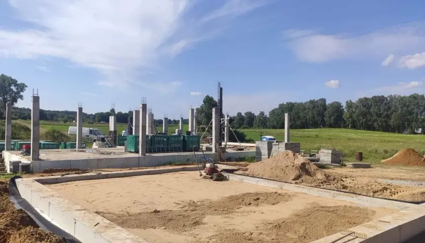 Budowa domu w toku. Betonowe słupy stoją na fundamencie, a w tle widać maszyny do zagęszczania gruntu i stosy piasku.