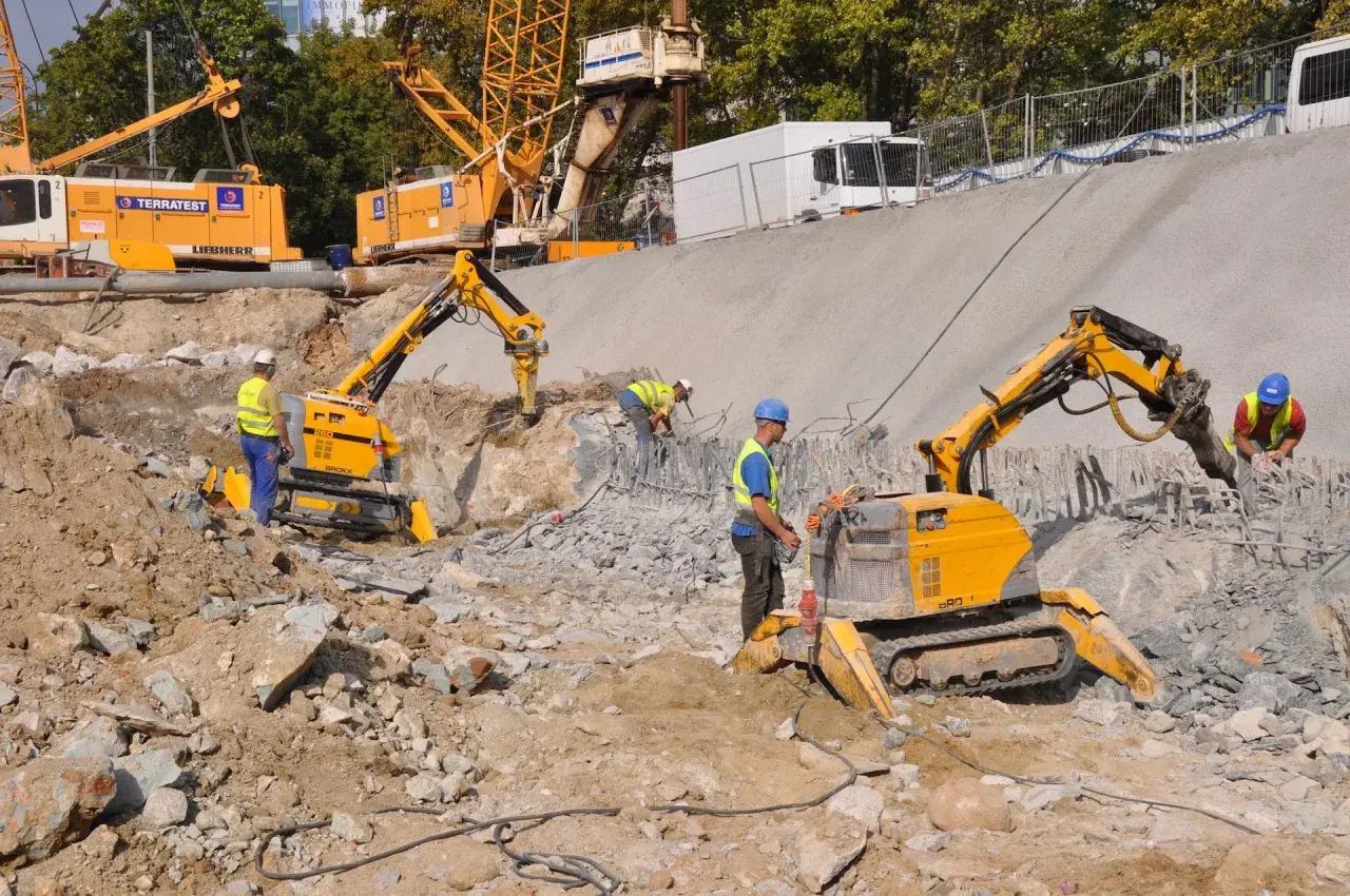 Na placu budowy pracują roboty wyburzeniowe, które kruszą beton. W tle widać koparki i dźwigi.