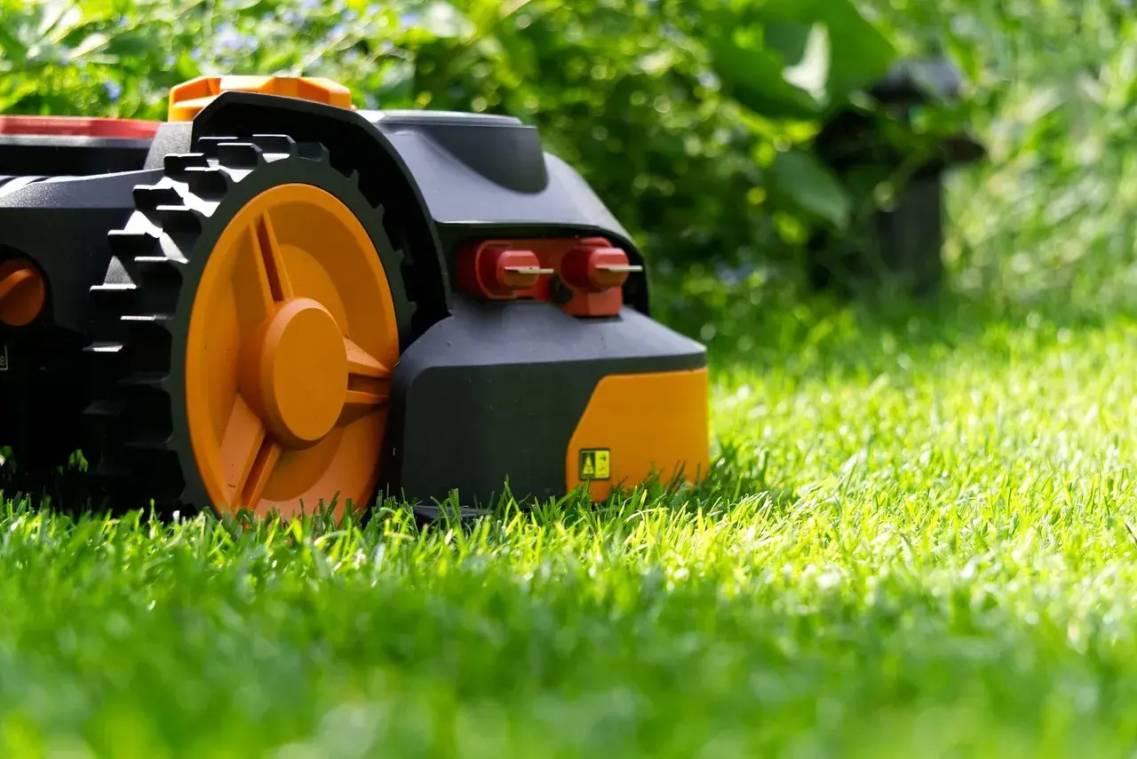 Robot koszący z pomarańczowymi kołami porusza się po soczystej trawie, pokazując, jak działa robot koszący.