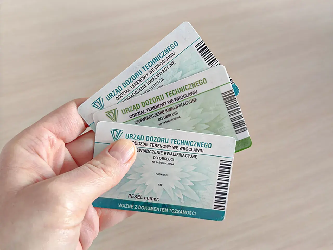 Ręka trzyma kilka zaświadczeń kwalifikacyjnych Urzędu Dozoru Technicznego, potwierdzających uprawnienia na zwyżki.