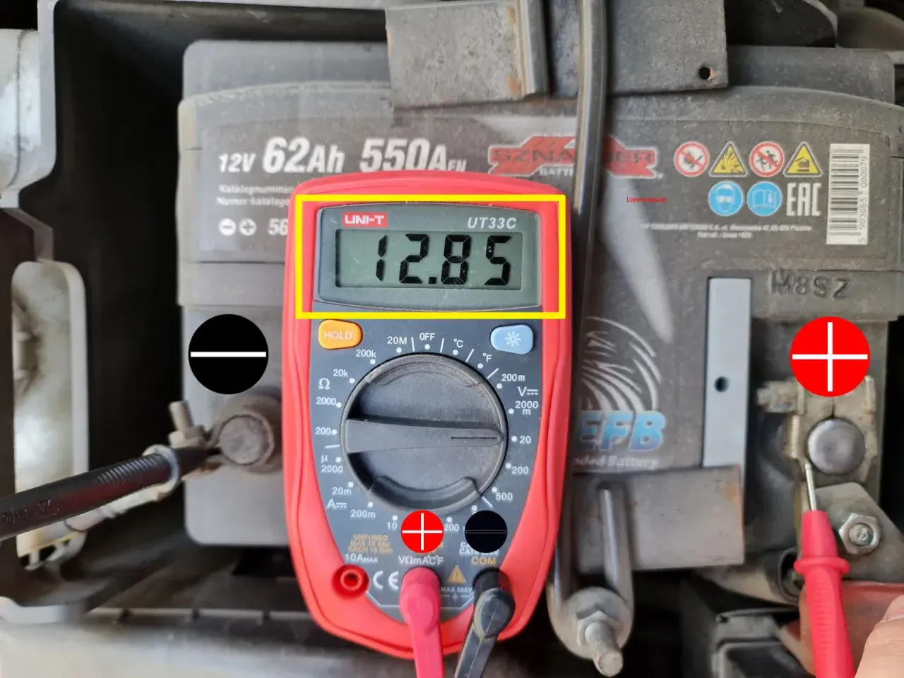 Multimetr pokazuje 12.85V, co jest wynikiem pomiaru napięcia elektrycznego akumulatora samochodowego.