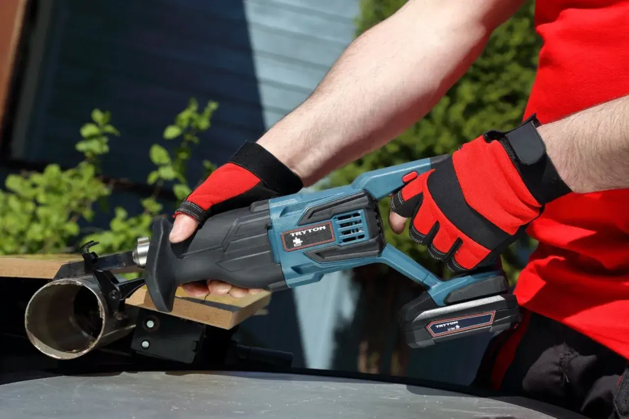 Ręce w czerwonych rękawicach trzymają piłę szablastą Tryton. To nie Black+Decker, ale równie solidne narzędzie do cięcia.