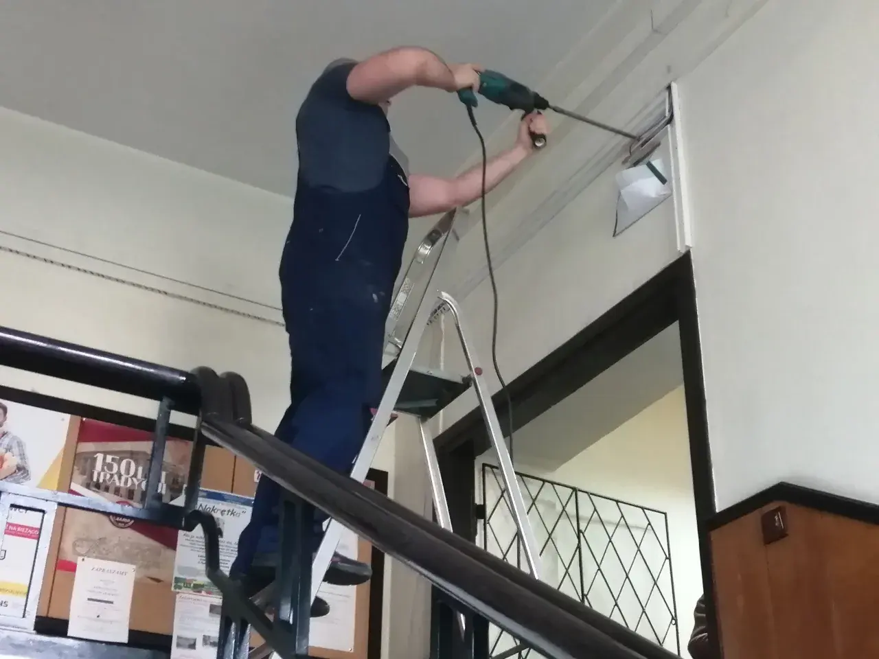 Mężczyzna w ogrodniczkach stoi na drabinie, wiercąc w ścianie. Trwa budowa drabiny, która ułatwia mu pracę.