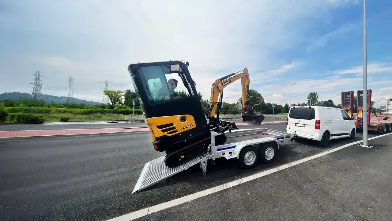 Mini koparka Sany SY18C na przyczepie, gotowa do transportu. Obok biały bus i inne przyczepy.