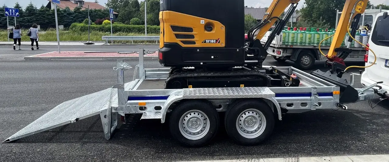 Żółta minikoparka na przyczepie, gotowa do transportu. Jej waga jest znacząca, ale przyczepa jest solidna.
