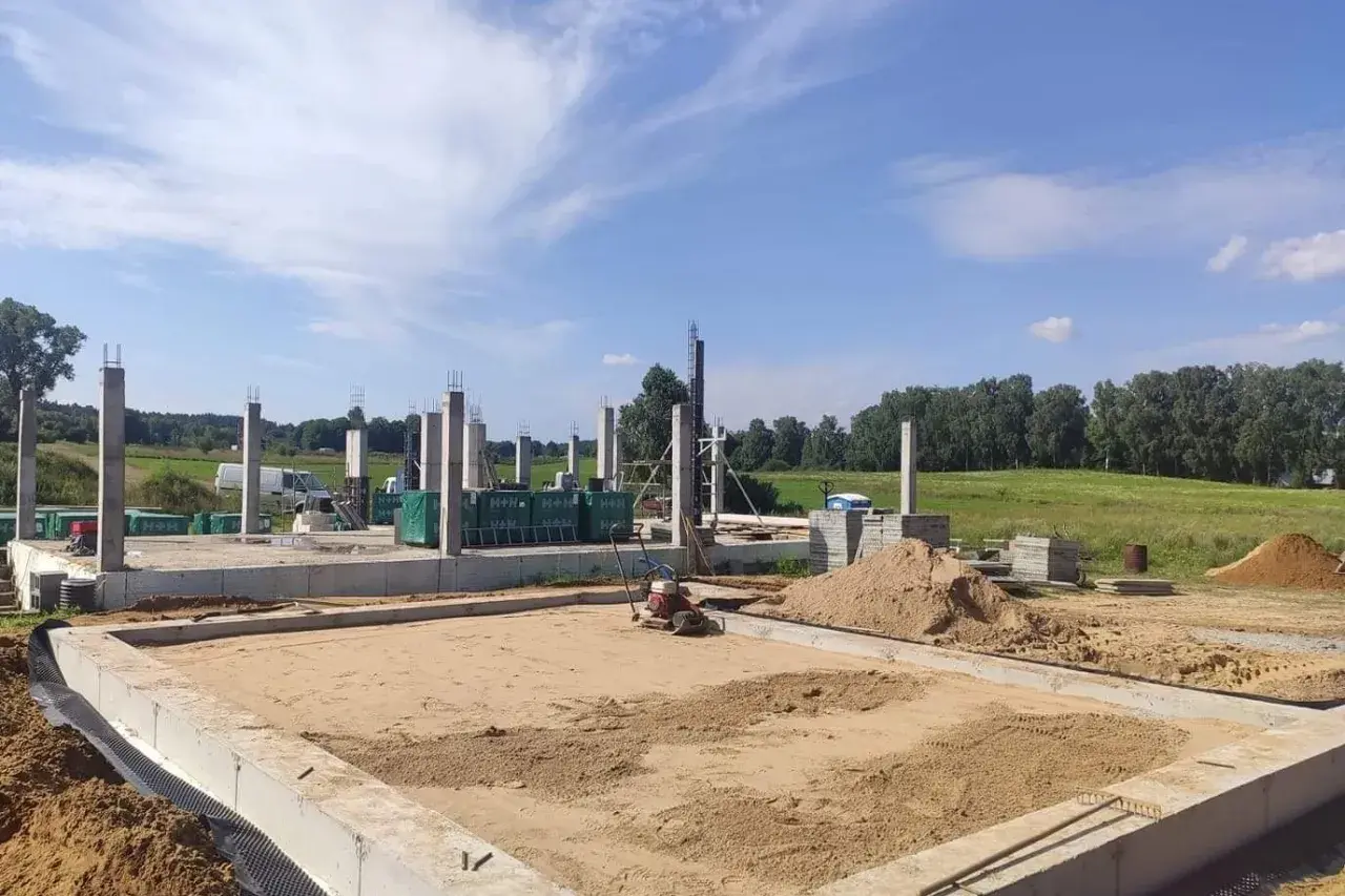 Budowa domu w toku. Betonowe słupy stoją na fundamencie, a w tle widać maszyny do zagęszczania gruntu i stosy piasku.
